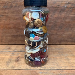 May include: A clear plastic jar with a black ribbed screw-top lid, filled with a variety of assorted buttons in different shapes, sizes, and colors, including metallic gold, brown, blue, pink, and white, with a small white label on the side.