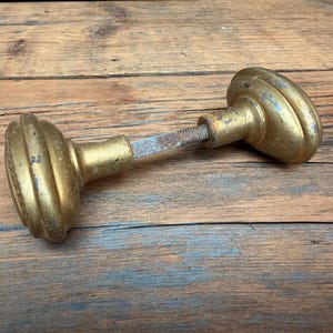 May include: A pair of vintage brass door knobs connected by a central metal spindle. The knobs feature a rounded, tiered design with a weathered, tarnished gold-toned finish showing signs of age and wear across the metallic surfaces.