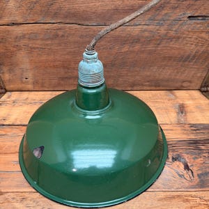 May include: A vintage-style dark green enameled metal pendant light shade with a dome shape. The fixture features a weathered, light-colored metal socket at the top with a short, frayed, brown fabric-covered electrical cord extending upward.