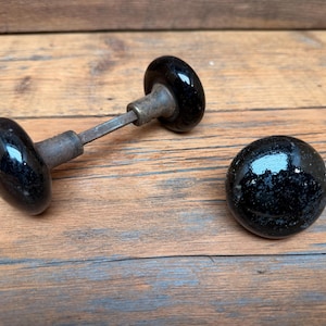 May include: Two round, glossy black door knobs with a smooth, reflective surface. One knob is attached to a square metal spindle and a cylindrical metal base, while the other knob is detached and sits separately as a single round piece.