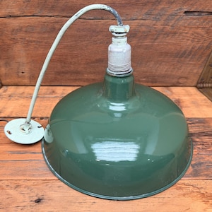 May include: A dark green enameled metal pendant light fixture with a wide, dome-shaped shade. The lamp features a central socket, a curved light-colored electrical cord, and a small circular mounting plate at the end of the cord.