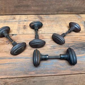 May include: Four dark brown, oval-shaped door knobs with a beaded edge detail. Each knob is attached to a central metal spindle, forming a complete door handle assembly. The knobs feature a smooth, rounded surface with a dark, weathered finish.