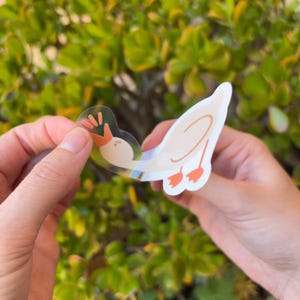 May include: A clear sticker of a cartoon goose with orange feet and a beak. The goose is looking to the left and has its wings spread out. The sticker is being peeled off of a white backing.