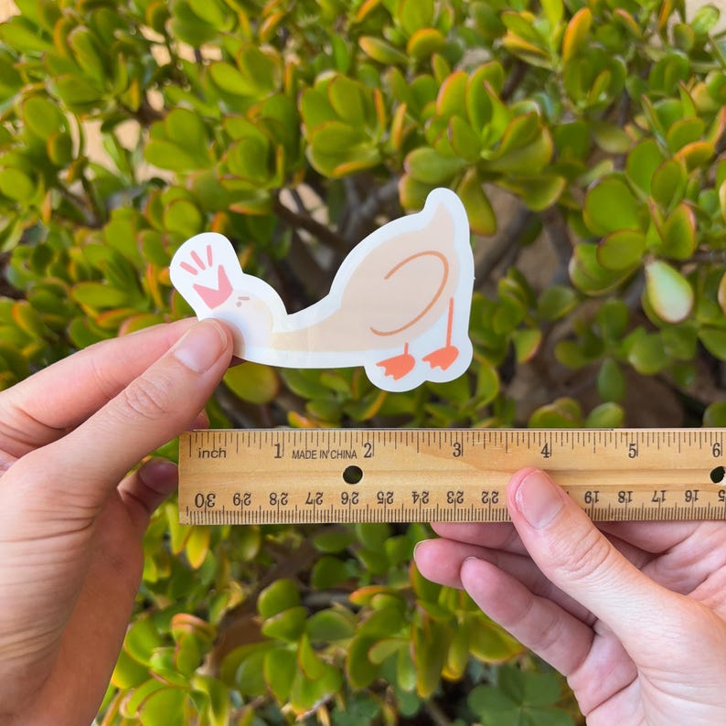 May include: A sticker of a cartoon duck with a pink handprint on its head. The sticker is about 5 cm long and is being held up next to a ruler.
