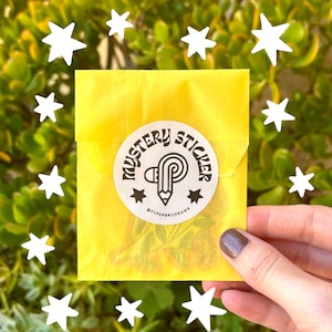 K&ouml;nnte beinhalten: Ein gelber Papierumschlag mit einem runden Aufkleber mit der Aufschrift "MYSTERY STICKER". Der Aufkleber zeigt einen Bleistift und Sterne. Wei&szlig;e Sterne umgeben den Umschlag, vor einem Hintergrund aus gr&uuml;nen Bl&auml;ttern.