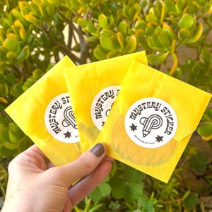 K&ouml;nnte beinhalten: Drei gelbe Papierumschl&auml;ge, jeweils mit einem wei&szlig;en Aufkleber mit der Aufschrift "Mystery Sticker" mit einem Bleistift- und Sterndesign. Die Umschl&auml;ge werden vor einem Hintergrund aus gr&uuml;nem Laub gehalten.