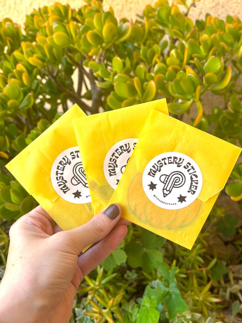 K&ouml;nnte beinhalten: Drei gelbe Papierumschl&auml;ge, jeweils mit einem wei&szlig;en Aufkleber mit der Aufschrift "Mystery Sticker" mit einem Bleistift- und Sterndesign. Die Umschl&auml;ge werden vor einem Hintergrund aus gr&uuml;nem Laub gehalten.