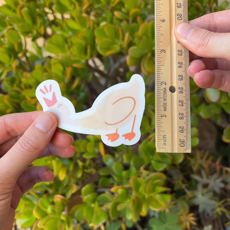 May include: A sticker of a light brown duck wearing a pink crown. The duck is about 5 cm tall and is holding its head up.