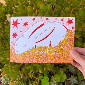 May include: A white bunny with red outlines is sleeping in a field of yellow flowers with red outlines. The background is pink with red stars.