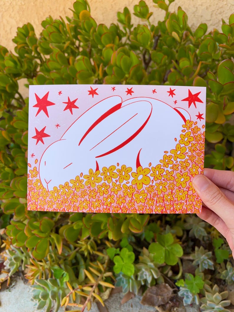May include: A white bunny with red outlines is sleeping in a field of yellow flowers with red outlines. The background is pink with red stars.