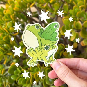May include: A sticker featuring three cartoon frogs stacked on top of each other in shades of green and yellow. White star-shaped accents surround the sticker, set against a backdrop of green foliage.