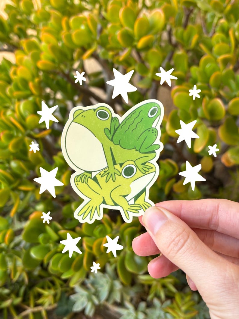 May include: A sticker featuring three cartoon frogs stacked on top of each other in shades of green and yellow. White star-shaped accents surround the sticker, set against a backdrop of green foliage.