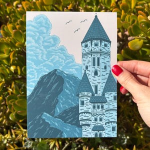 May include: A blue and white art print featuring a castle with multiple turrets, mountains, and clouds. Three birds fly in the sky above the castle. The print is held by a person with red nail polish.