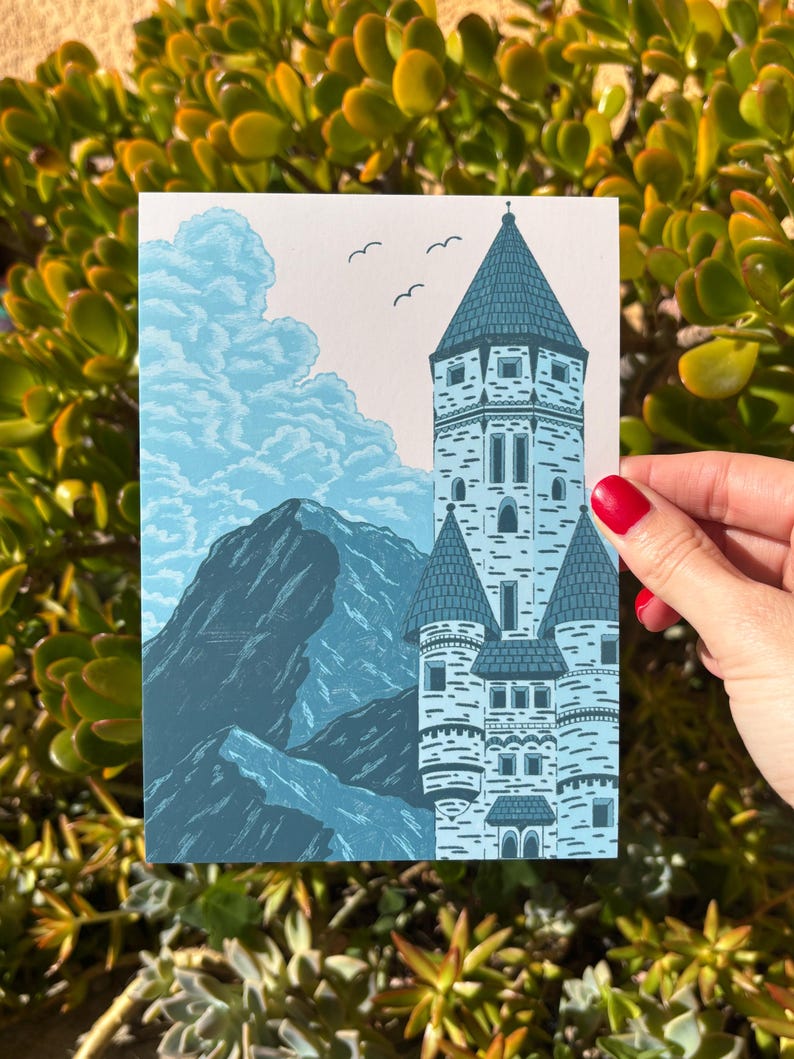 May include: A blue and white art print featuring a castle with multiple turrets, mountains, and clouds. Three birds fly in the sky above the castle. The print is held by a person with red nail polish.