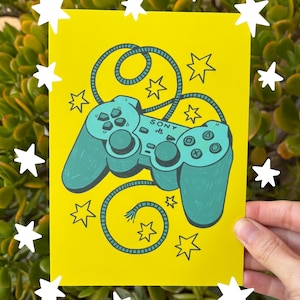 May include: A yellow card featuring a teal-colored illustration of a classic video game controller with the word "SONY" on it. The card is decorated with teal stars and a teal cord. White star stickers surround the card.