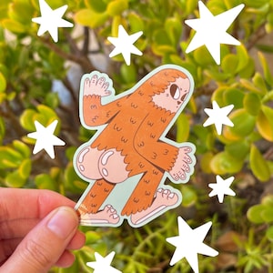 May include: A sticker featuring a cartoon Sasquatch with brown fur, light pink skin, and a light blue background. White star stickers are scattered around the image. The sticker is held by a hand in front of a green leafy background.