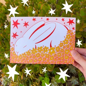 May include: A white bunny with red outlines is nestled in a field of yellow and orange flowers. The background is a pink gradient with white stars.