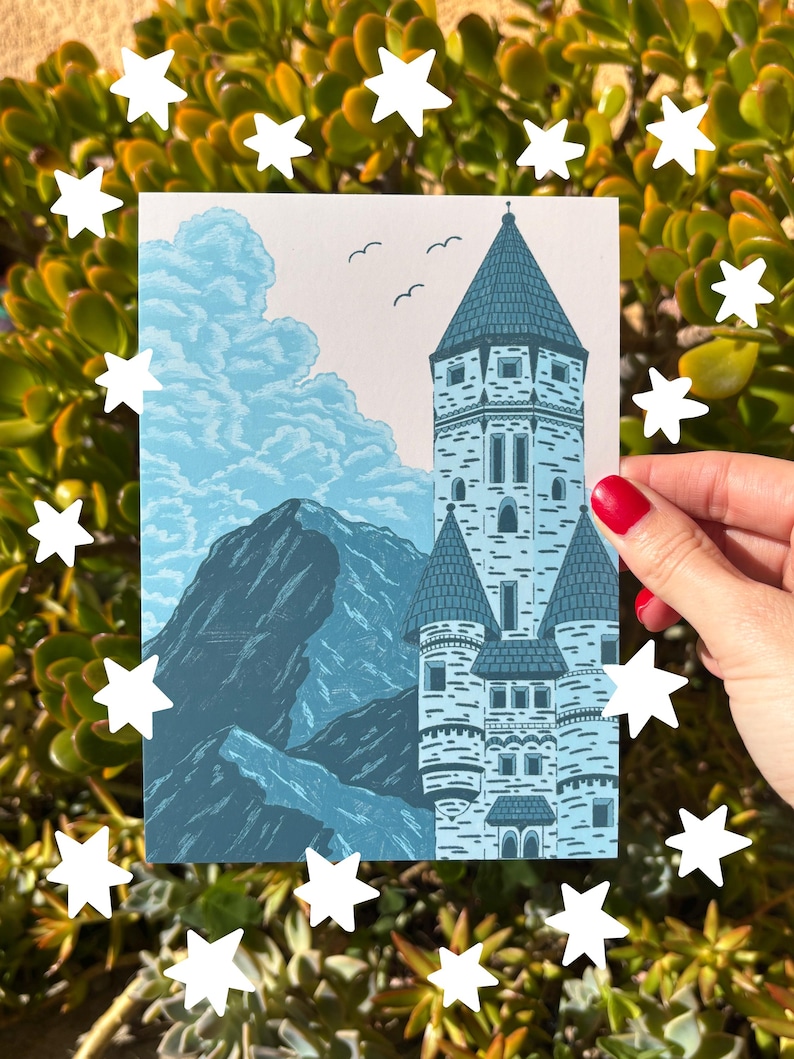 May include: A blue and white print of a castle with a mountain range and clouds in the background. The castle has multiple turrets and a tall central tower. Three birds fly above the castle. The print is surrounded by white star cutouts.