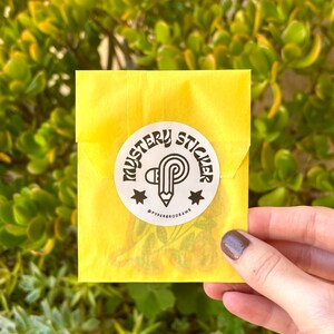 K&ouml;nnte beinhalten: Ein gelber Papierumschlag mit einem wei&szlig;en runden Aufkleber mit der Aufschrift "Mystery Sticker" in einer stilisierten Schriftart. Der Aufkleber zeigt einen Bleistift und den Text "@pyperrodraws". Der Umschlag wird vor einem gr&uuml;nen, bl&auml;ttrigen Hintergrund gehalten.
