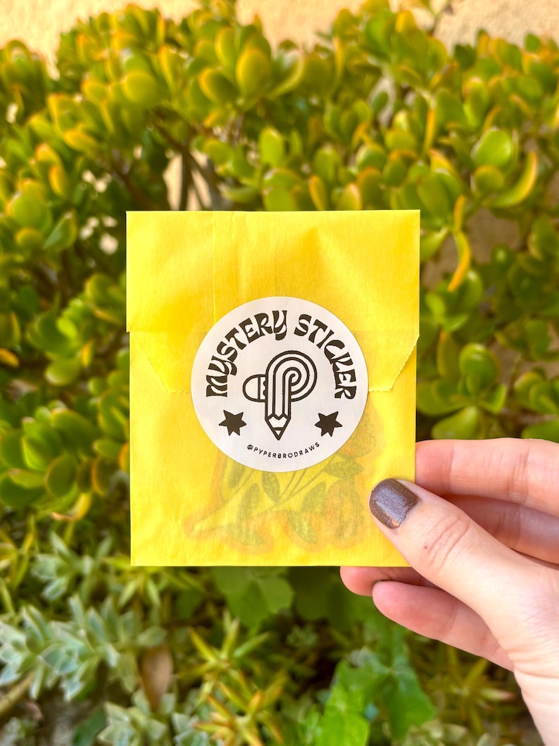 K&ouml;nnte beinhalten: Ein gelber Papierumschlag mit einem wei&szlig;en runden Aufkleber mit der Aufschrift "Mystery Sticker" in einer stilisierten Schriftart. Der Aufkleber zeigt einen Bleistift und den Text "@pyperrodraws". Der Umschlag wird vor einem gr&uuml;nen, bl&auml;ttrigen Hintergrund gehalten.