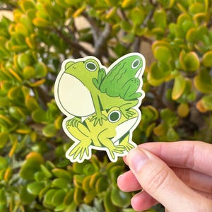 May include: A sticker featuring three cartoon frogs stacked on top of each other. The frogs are various shades of green with black eyes and white highlights. The sticker is held against a background of green foliage.