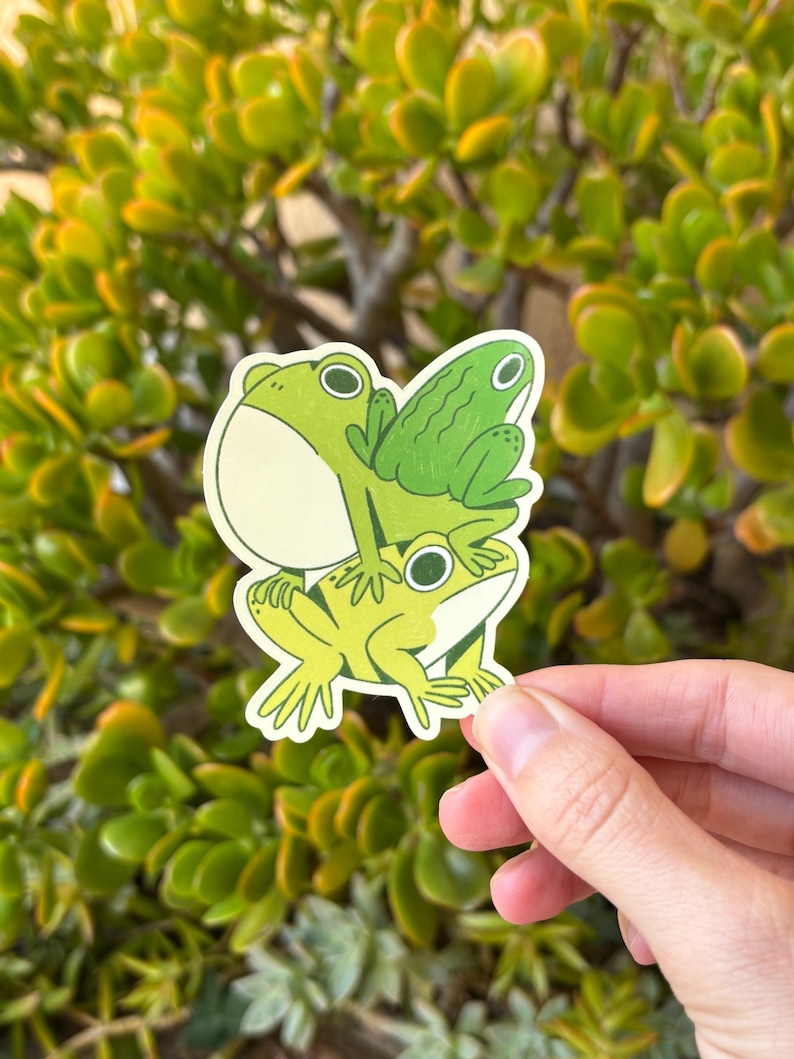 May include: A sticker featuring three cartoon frogs stacked on top of each other. The frogs are various shades of green with black eyes and white highlights. The sticker is held against a background of green foliage.