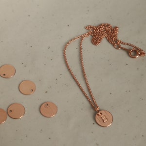 Peut inclure: Un collier en or rose avec un pendentif circulaire grav&eacute; de la lettre "B". La cha&icirc;ne et le pendentif du collier sont pr&eacute;sent&eacute;s sur une surface grise clair mouchet&eacute;e, avec plusieurs &eacute;tiquettes m&eacute;talliques circulaires vierges &agrave; proximit&eacute;.
