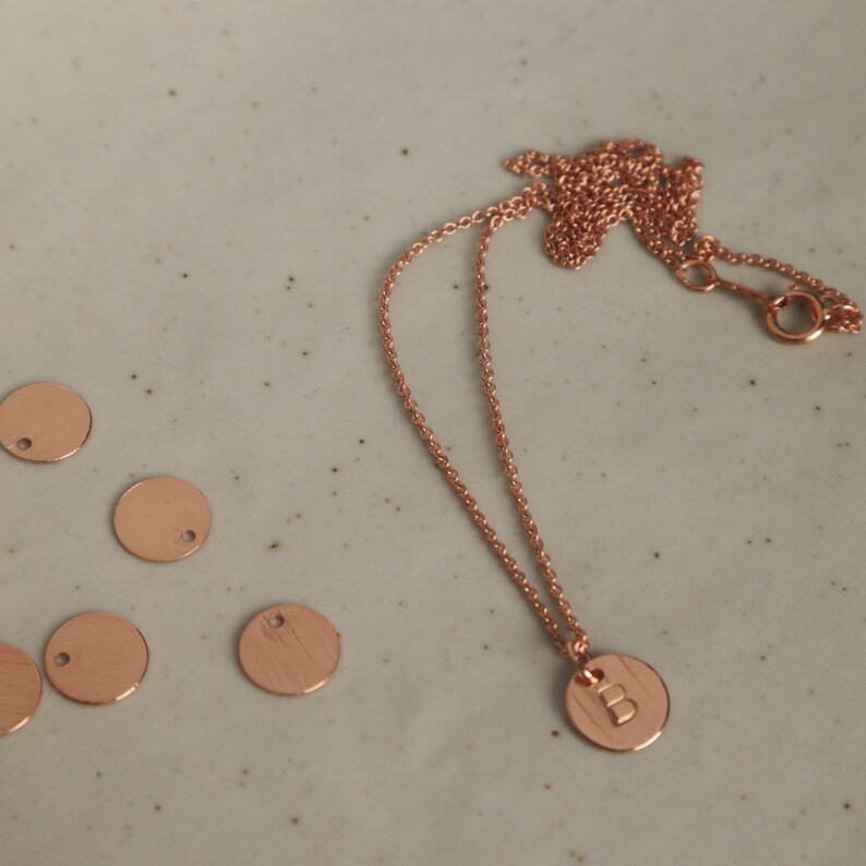 Peut inclure: Un collier en or rose avec un pendentif circulaire grav&eacute; de la lettre "B". La cha&icirc;ne et le pendentif du collier sont pr&eacute;sent&eacute;s sur une surface grise clair mouchet&eacute;e, avec plusieurs &eacute;tiquettes m&eacute;talliques circulaires vierges &agrave; proximit&eacute;.
