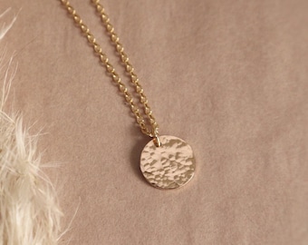 Può includere: Una collana color oro caratterizzata da una catenina sottile e un piccolo ciondolo rotondo con una superficie martellata e testurizzata. Il ciondolo circolare riflette la luce sulla sua finitura irregolare e increspata, appeso a una catena a maglie fini.