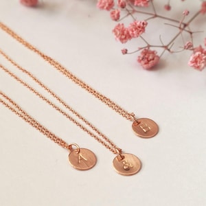 Peut inclure: Trois colliers d&eacute;licats en or rose avec des pendentifs circulaires, chacun grav&eacute; d'une seule lettre. Les colliers sont pr&eacute;sent&eacute;s sur une surface blanche, avec des fleurs roses en arri&egrave;re-plan. Les pendentifs mesurent environ 1,3 cm de diam&egrave;tre.