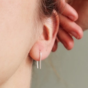 Könnte beinhalten: Ein Paar silberne Minimalistische Ohrringe mit einem einfachen, geraden Design. Die Ohrringe sind im Ohrläppchen einer Person zu sehen.