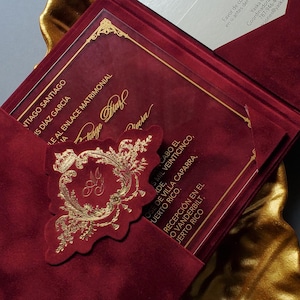May include: Burgundy velvet wedding invitation with gold foil accents. The invitation features a gold emblem and text, presented in a gold leaf-shaped dish. Includes a separate card with text.