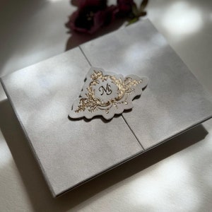 May include: A white invitation card with a gold embossed design featuring the letter "M" in a circle. The card is folded in half and has a decorative edge. The card is surrounded by flowers.