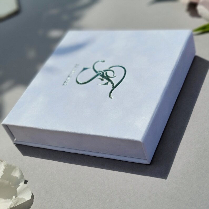Puede incluir: Caja blanca con un dise&ntilde;o en relieve plateado de las letras "S&S" en la parte superior. La caja est&aacute; cerrada.