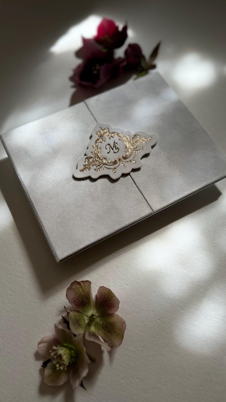 May include: A white invitation card with a gold embossed design featuring the letter "M" in a circle. The card is folded in half and has a decorative edge. The card is surrounded by flowers.