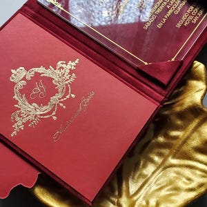 May include: Red wedding invitation with gold foil accents, featuring the names "Andrea and Jesus". The invitation is inside a red velvet folder, with a clear overlay. The invitation is placed on a gold leaf-shaped tray.