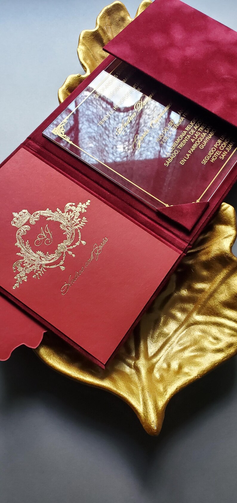May include: Red wedding invitation with gold foil accents, featuring the names "Andrea and Jesus". The invitation is inside a red velvet folder, with a clear overlay. The invitation is placed on a gold leaf-shaped tray.