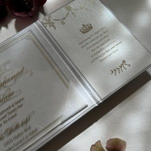 May include: A white wedding invitation with gold lettering and a floral design. The invitation reads "And among His Signet is that...that He created for you mater...from among yourselves...that you may dwell...in tranquillity with them...and He has put love and mercy...between your hearts". The invitation also includes the date, time, and location of the wedding.