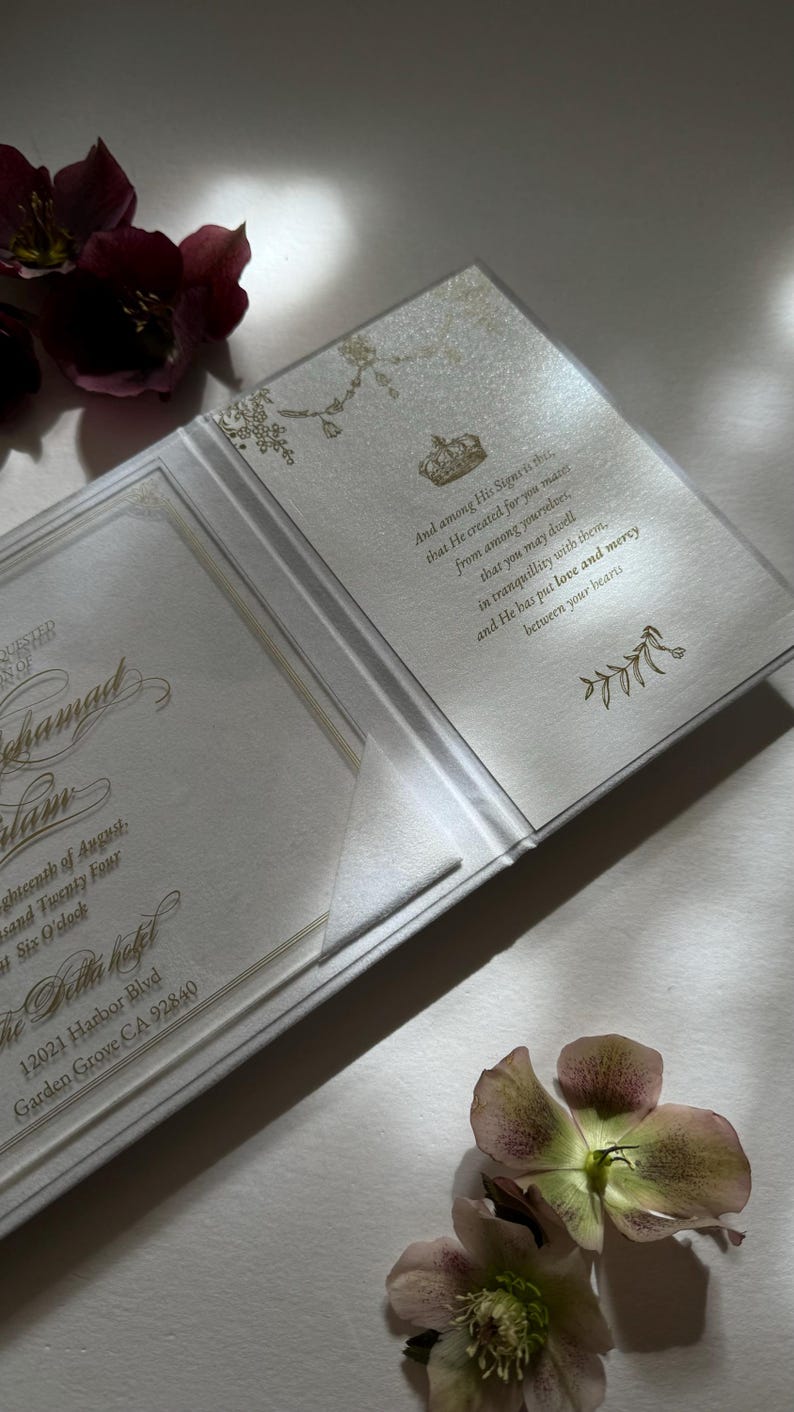May include: A white wedding invitation with gold lettering and a floral design. The invitation reads "And among His Signet is that...that He created for you mater...from among yourselves...that you may dwell...in tranquillity with them...and He has put love and mercy...between your hearts". The invitation also includes the date, time, and location of the wedding.