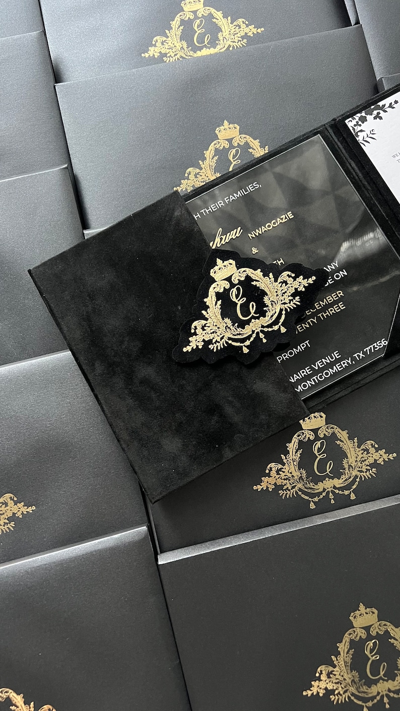 May include: Black velvet invitation with a clear acrylic insert. The insert features gold lettering with the names "Kuru & NWAOGAZIE" and the date "December Twenty Three". The invitation also features a gold crest with the initials "E & E" and a crown.