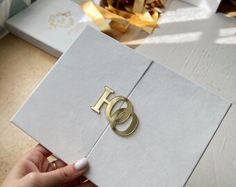 May include: White wedding invitation with a gold foil design. The invitation is folded in half and has two interlocking rings on the front. The rings are made of gold metal and have a shiny finish.