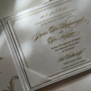 May include: A white wedding invitation with gold lettering. The invitation announces the wedding of Juris Dr. Mhamad and Dr. Salam. The wedding will take place on Sunday, the eighteenth of August, Two Thousand Twenty Four at Six O'clock at The Bella Hotel, 12021 Harbor Blvd, Garden Grove CA 92840.