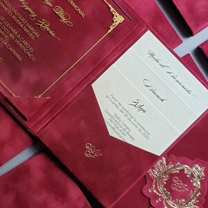 May include: Burgundy wedding invitations with gold foil accents. The invitations feature elegant script and decorative embellishments. The design includes a pocket-style invitation with inserts and a matching enclosure card.