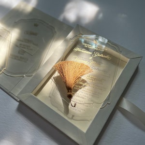 Peut inclure: Une invitation de mariage en acrylique transparente avec des lettres dorées. L'invitation est dans une boîte blanche avec un pompon doré. Le texte sur l'invitation est "God has led two lives to take one path" et "Holumwani & Gabrielle Vincento Gabrielle" suivi de la date, de l'heure et du lieu du mariage. L'invitation comprend également le code vestimentaire, "Black Tie."