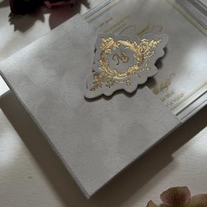May include: A white envelope with a gold foil monogram and floral design. The envelope is open and the inside is visible. The envelope is surrounded by pink and burgundy flowers.