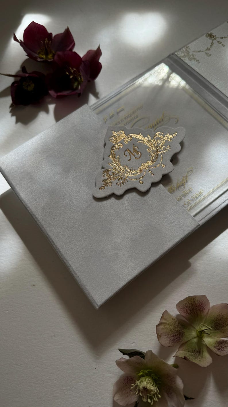 May include: A white envelope with a gold foil monogram and floral design. The envelope is open and the inside is visible. The envelope is surrounded by pink and burgundy flowers.