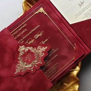 May include: Burgundy wedding invitation with gold accents, featuring a decorative emblem and text. The invitation is displayed on a gold leaf-shaped tray, creating an elegant presentation.