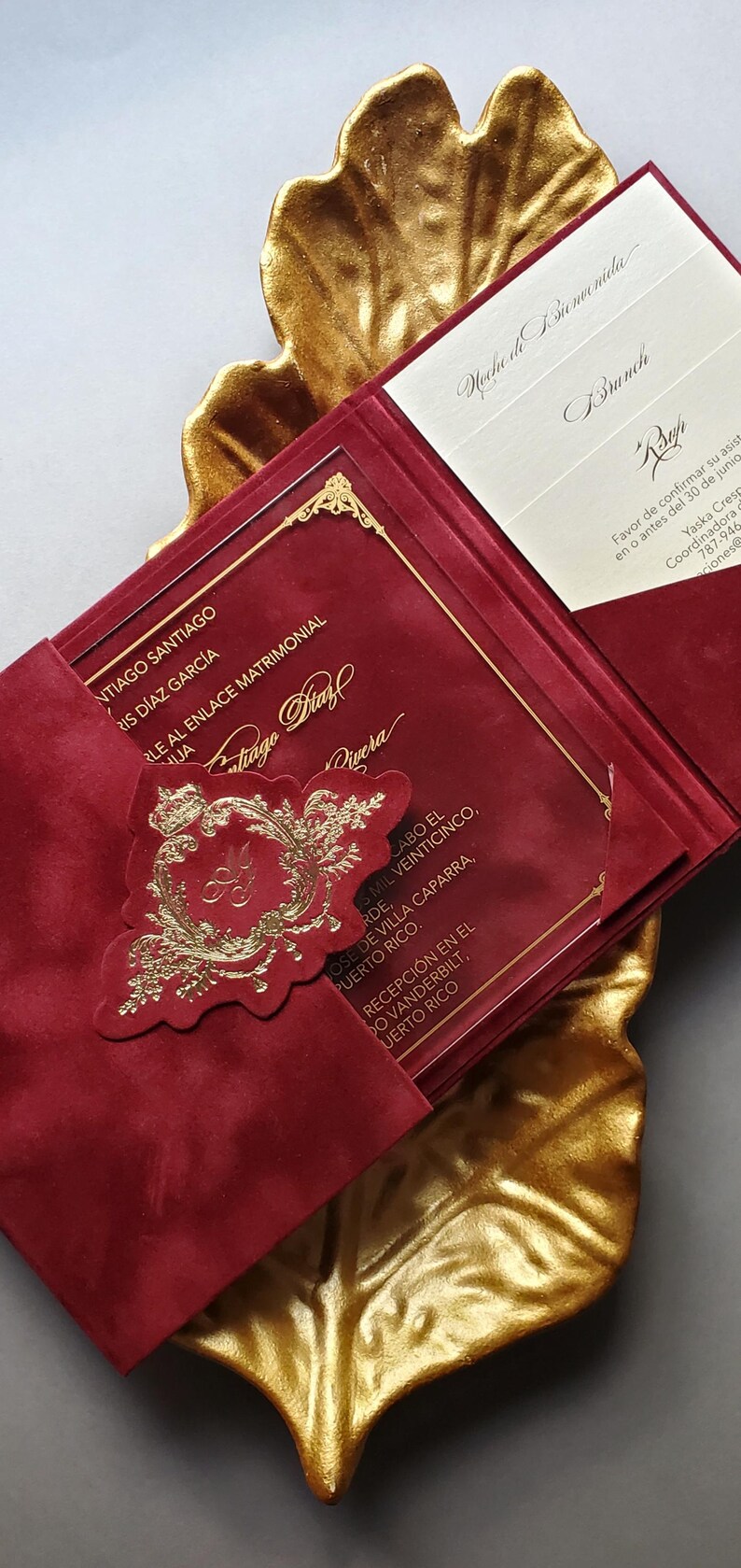 May include: Burgundy wedding invitation with gold accents, featuring a decorative emblem and text. The invitation is displayed on a gold leaf-shaped tray, creating an elegant presentation.