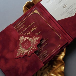 May include: Burgundy wedding invitation with gold accents, featuring a gold embossed emblem and elegant script. The invitation is presented on a gold leaf-shaped tray, creating a luxurious aesthetic.