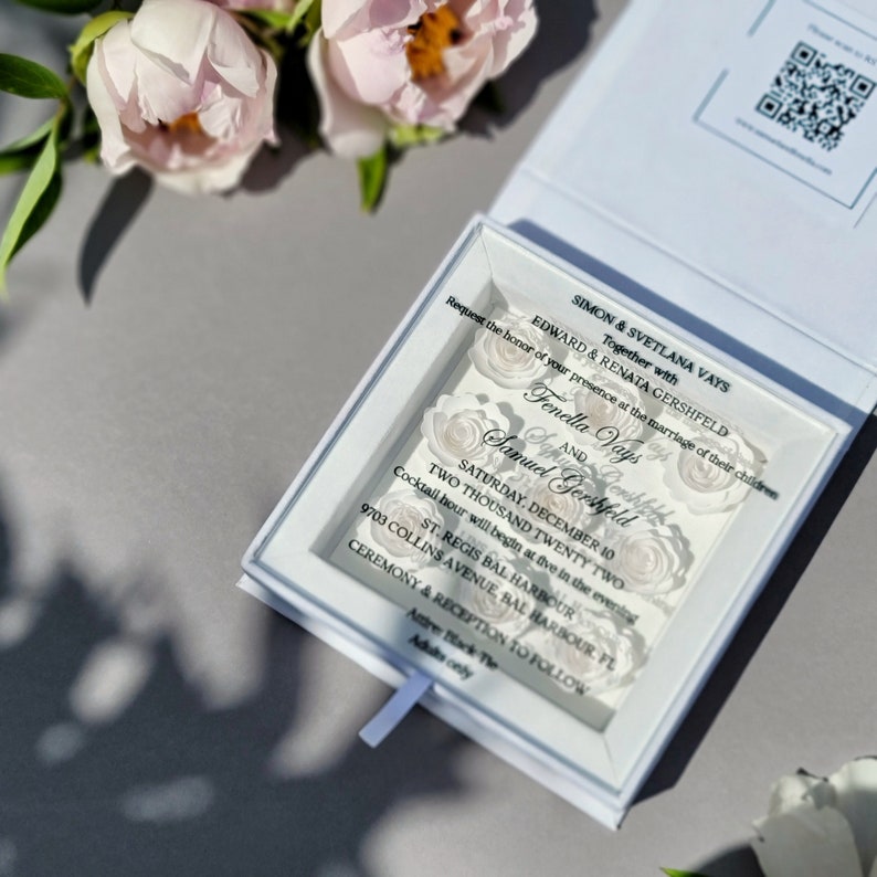Puede incluir: Una caja blanca con una ventana de acr&iacute;lico transparente que muestra una invitaci&oacute;n de boda. La invitaci&oacute;n presenta un dise&ntilde;o floral con el texto "Simon & Svetlana Vays Junto con Edward & Renata Gershfeld Solicitamos el honor de su presencia en el matrimonio de sus hijos Fenella Vays y Samuel Gershfeld S&aacute;bado, 10 de diciembre Dos mil veintid&oacute;s St. Regis Bal Harbour 9703 Collins Avenue, Bal Harbour FL Ceremonia y recepci&oacute;n a continuaci&oacute;n Solo adultos"